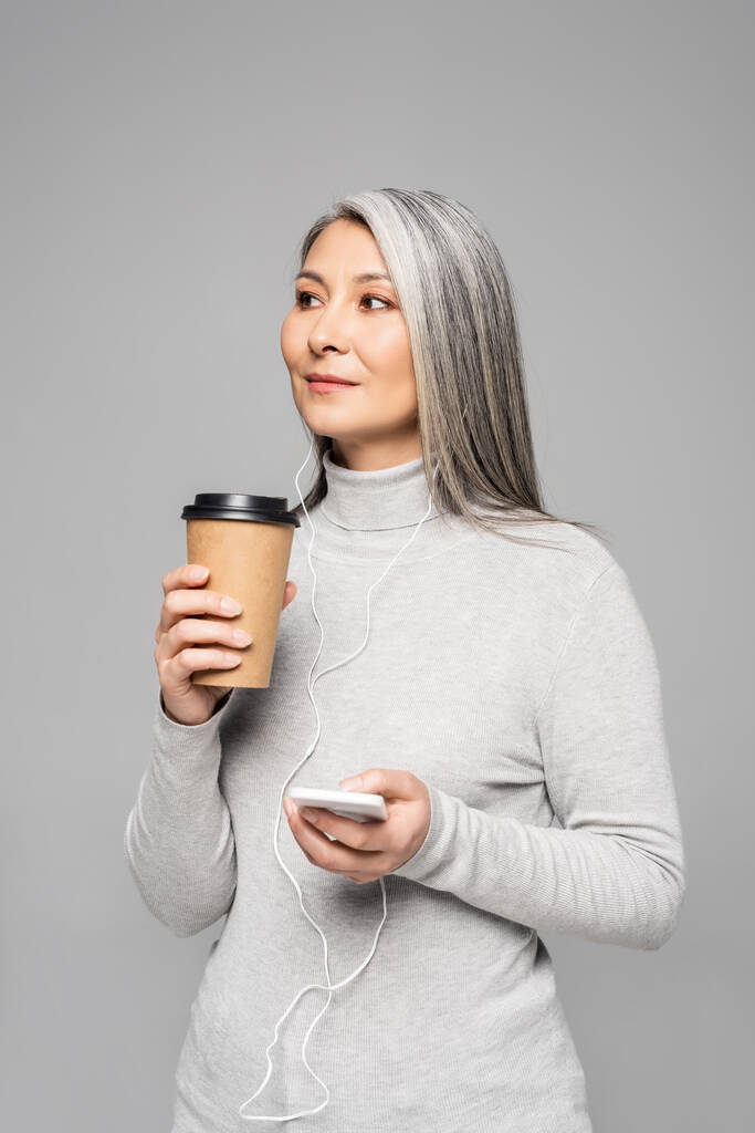 schöne asiatische Frau im Rollkragen mit Kaffee zum Musikhören mit Kopfhörern und Smartphone isoliert auf grau - Foto, Bild
