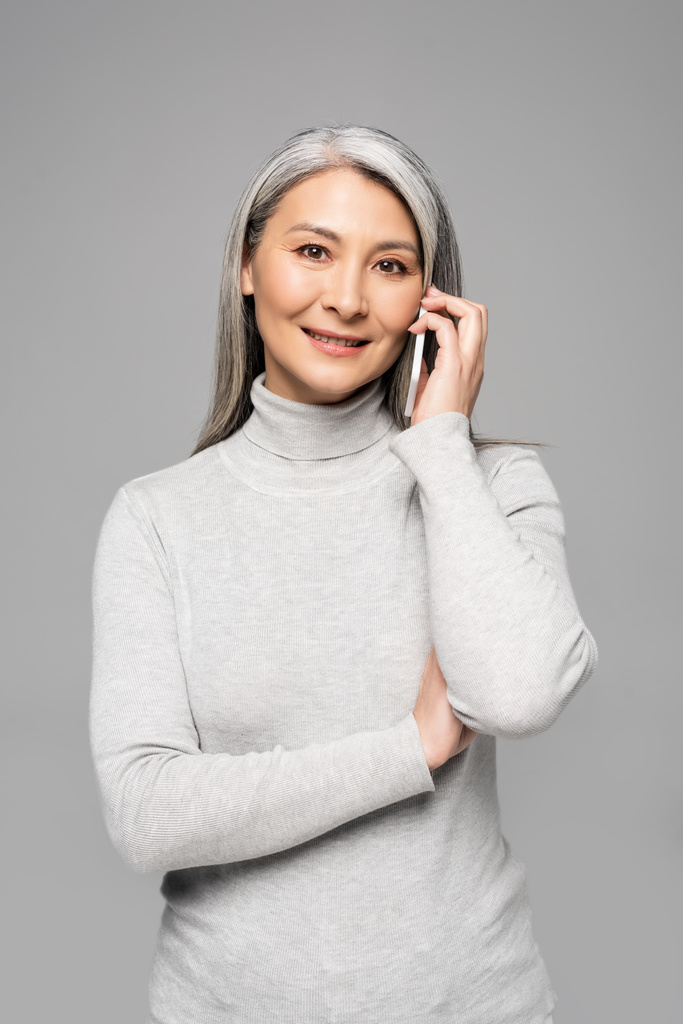 šťastná asijská žena v roláku s šedými vlasy mluví na smartphone izolované na šedé - Fotografie, Obrázek