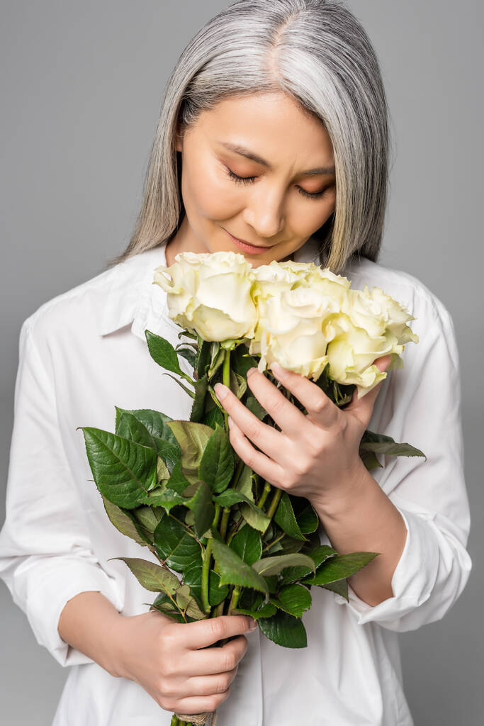 adulto concurso asiático mulher com cabelos grisalhos e olhos fechados segurando buquê de rosas brancas isolado no cinza
 - Foto, Imagem