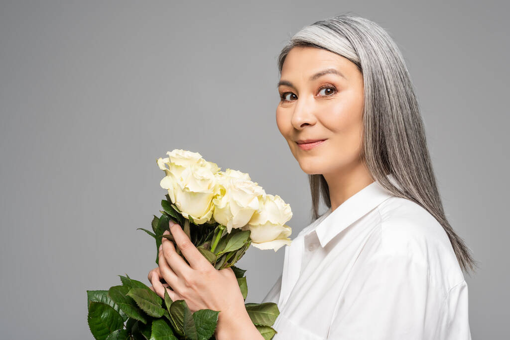 Erwachsene lächelnde asiatische Frau mit grauen Haaren, die einen Strauß weißer Rosen auf grau isoliert hält - Foto, Bild