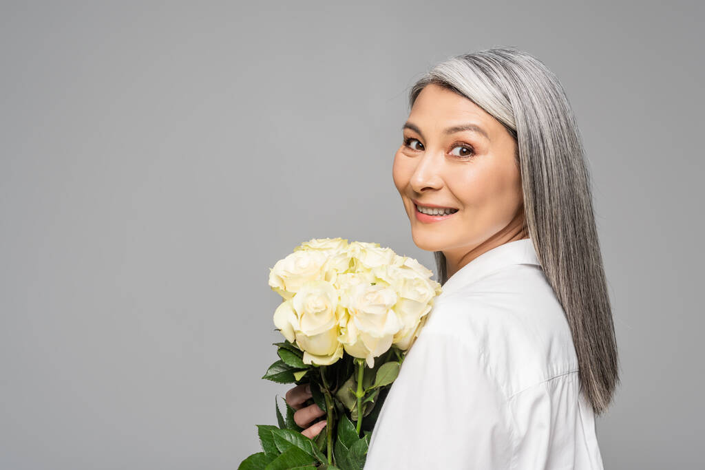 Gri saçlı, yetişkin, mutlu Asyalı kadın. Gri üzerinde izole edilmiş beyaz güller tutuyor. - Fotoğraf, Görsel
