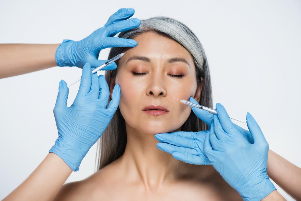 nahá asijská žena a lékaři v latexových rukavicích držící injekční stříkačky s kosmetickými injekcemi izolované na šedé - Fotografie, Obrázek