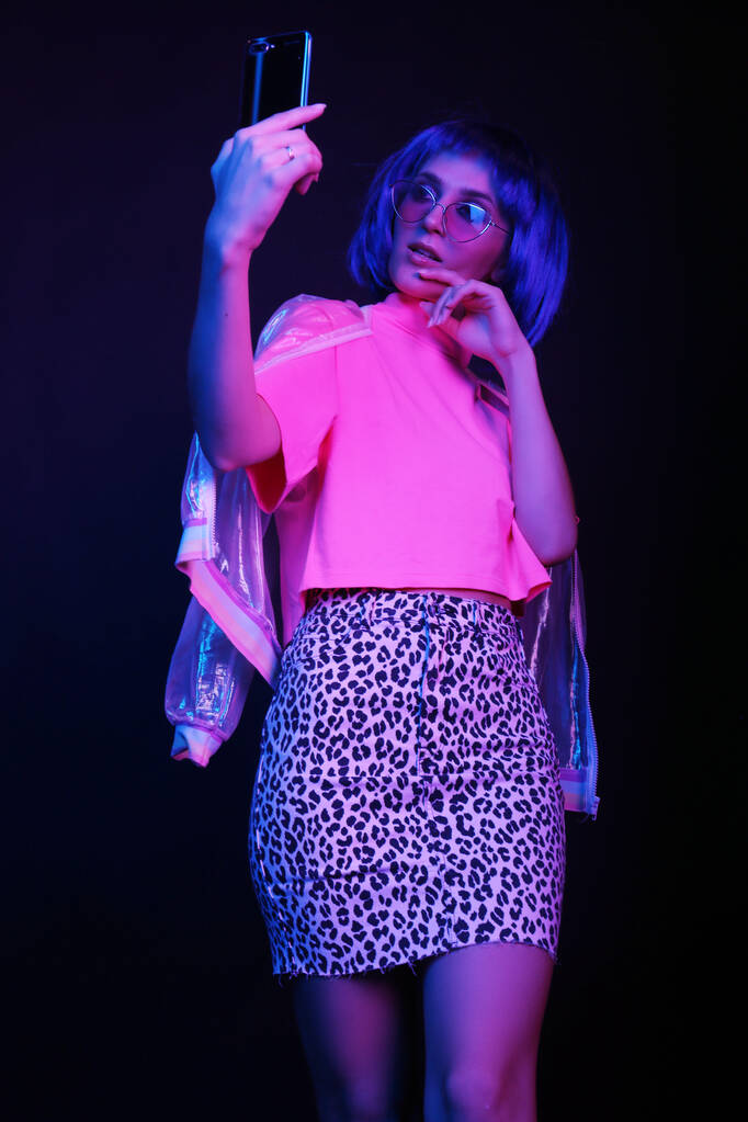 Modieuze jonge vrouw in een neon licht dansend in een nachtclub. Het meisje draagt een pruik, een bril en een glanzend, halo jasje, luipaard rok. Maakt een selfie op een smartphone. - Foto, afbeelding