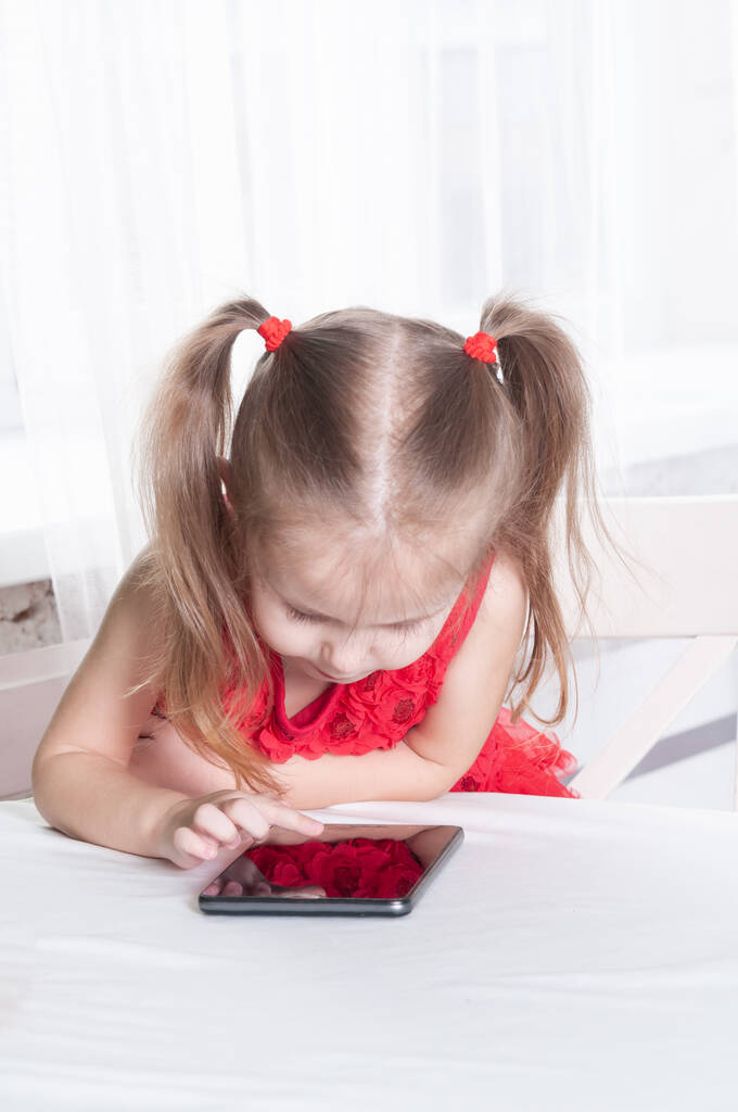 menina em um vestido vermelho senta-se a uma mesa e brinca com um tablet, com um sorriso no rosto
 - Foto, Imagem