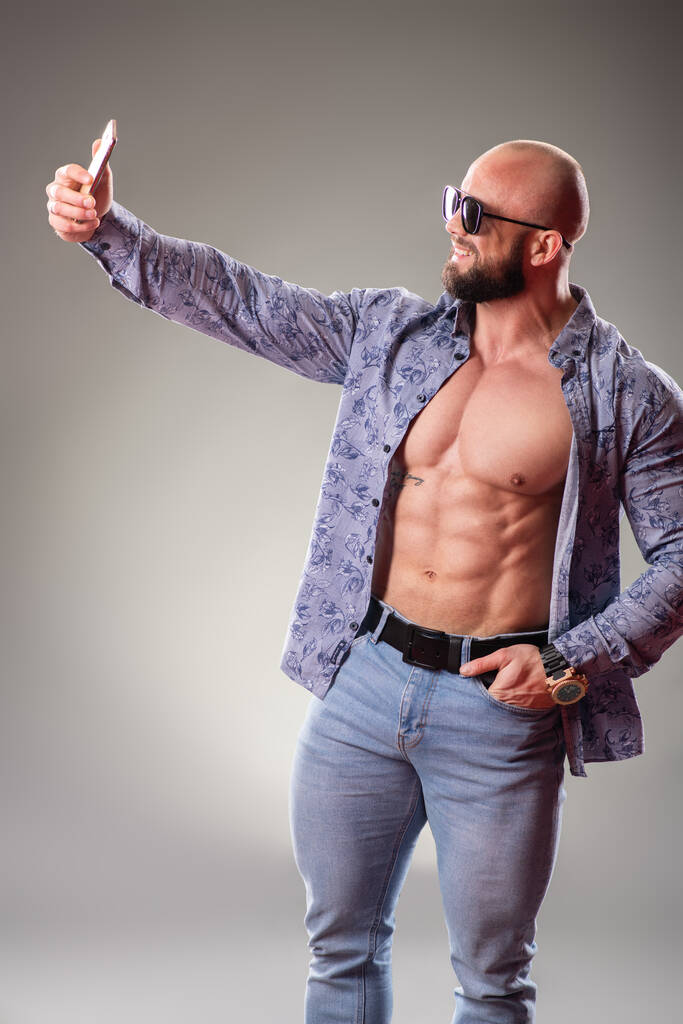 Sexy unbuttoned muscular bodybuider with sunglasses on the gray background. Selfie concept - Photo, Image