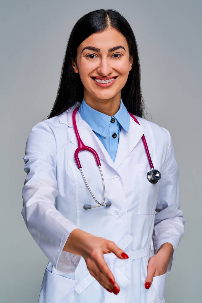 Γυναίκα με χαμόγελο με μελαχρινή μακριά μαλλιά σε ένα ιατρικό παλτό με κόκκινα νύχια κοιτάζοντας την κάμερα - Φωτογραφία, εικόνα