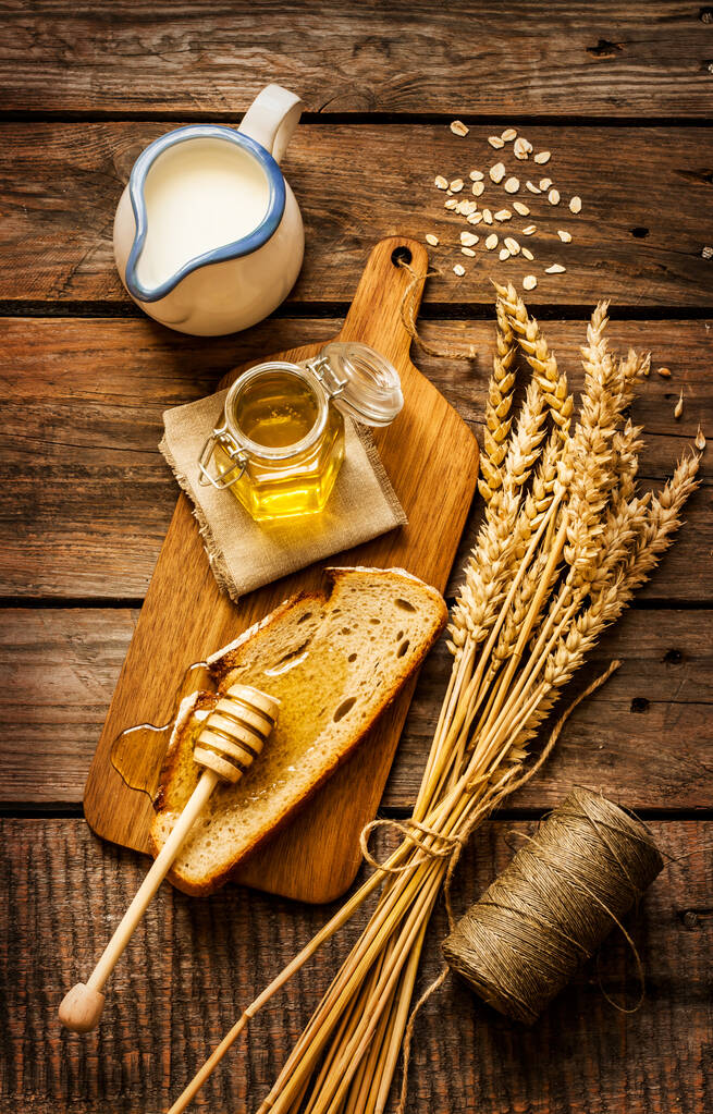 瓶の中の蜂蜜、上から古いヴィンテージの木の背景にパン、小麦、牛乳のスライス。田舎風または素朴なスタイルの朝食コンセプト. - 写真・画像