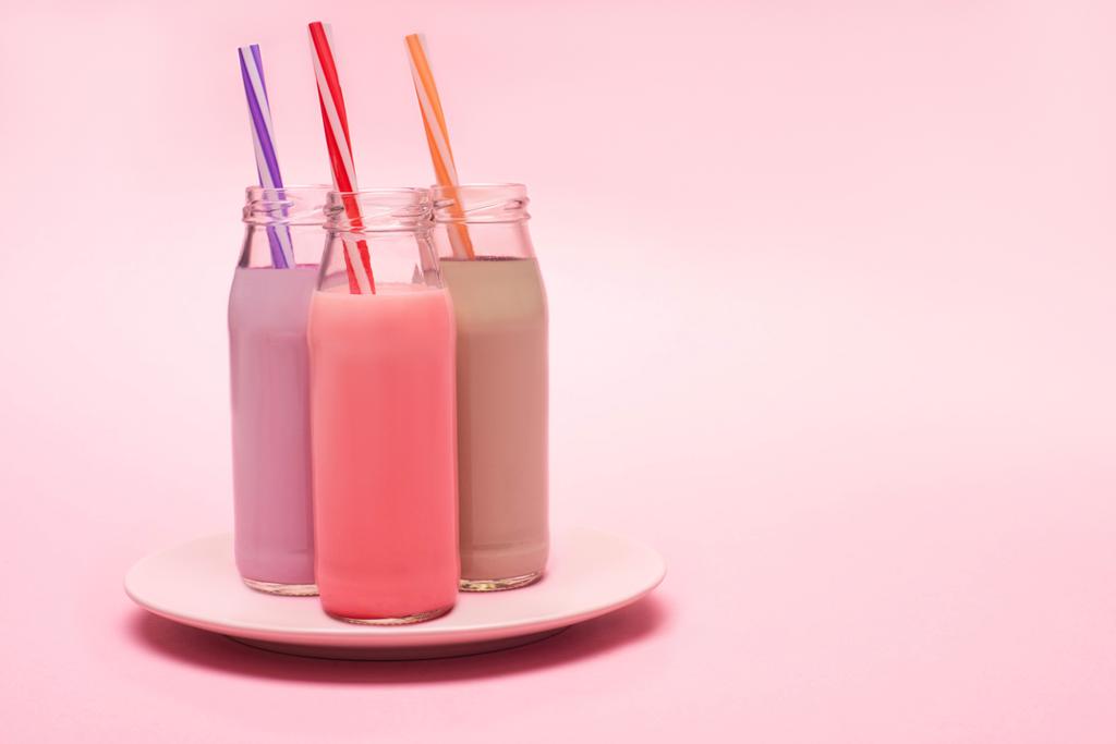 Bottles of berry, strawberry and chocolate milkshakes with drinking straws on plate on pink background - Photo, Image