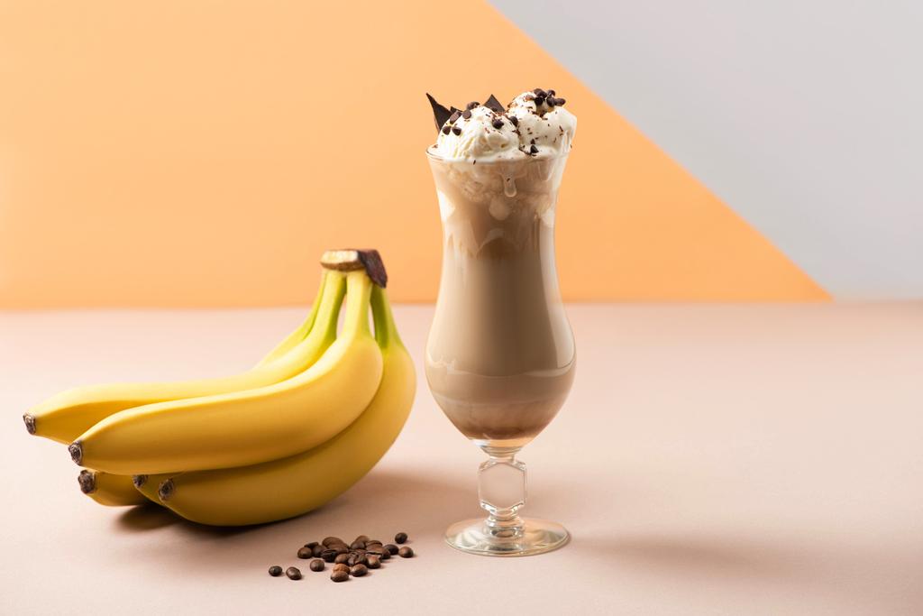Bicchiere di frullato con bocconcini di cioccolato vicino a banane e chicchi di caffè su beige, grigio e arancione
 - Foto, immagini
