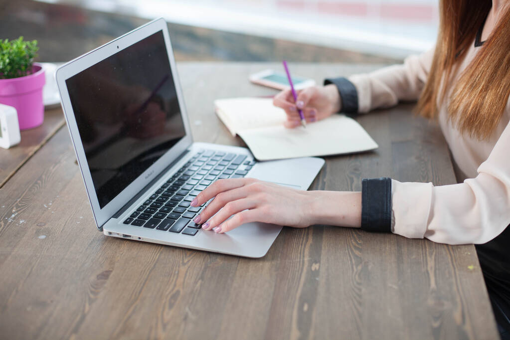 Schreibtisch mit Laptop, Tablet. In weiblichen Händen ein fuchsiafarbener Bleistift. Gelbe Blätter zum Schreiben, Bleistifte und eine Blume im Topf auf dem Tisch - Foto, Bild