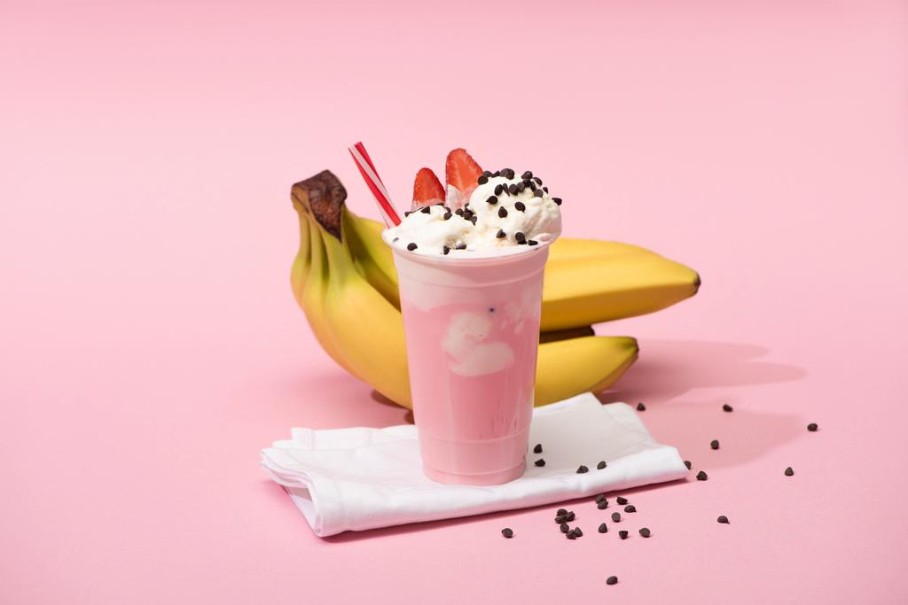Disposable cup of strawberry milkshake with chocolate chips on napkins near bananas on pink background - Photo, Image