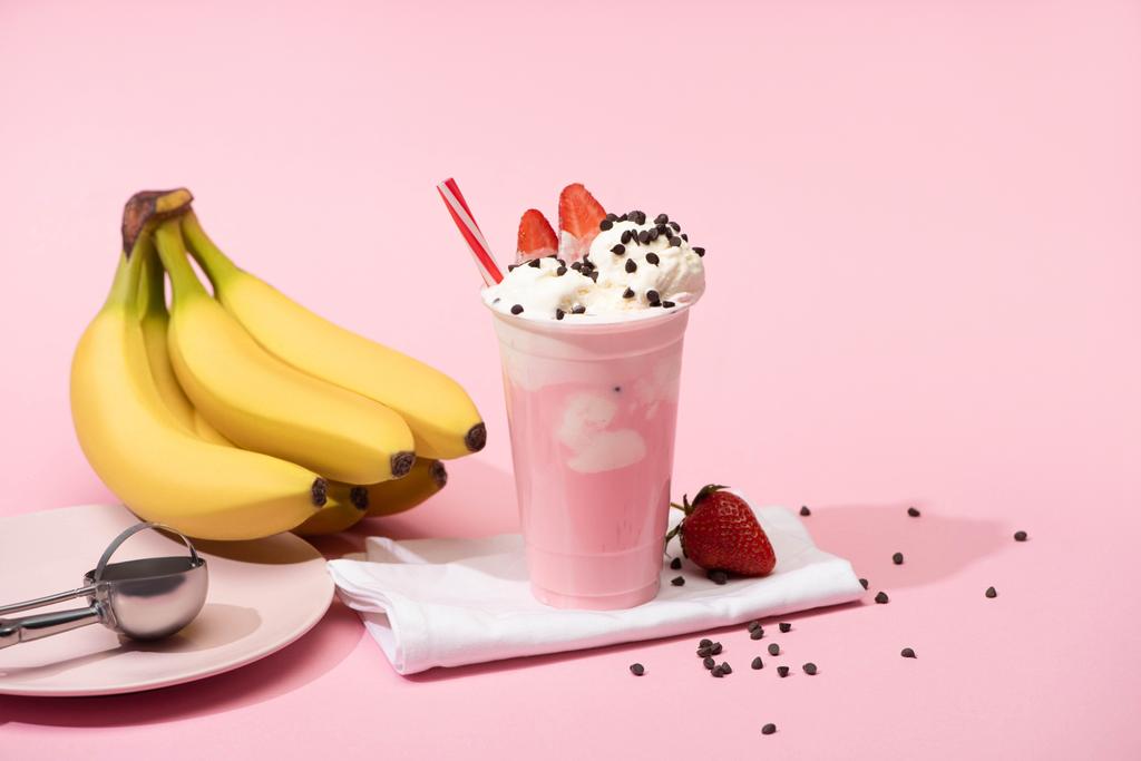 Disposable cup of milkshake with chocolate morsels and strawberries on napkins near bananas and plate with scoop on pink  - Photo, Image