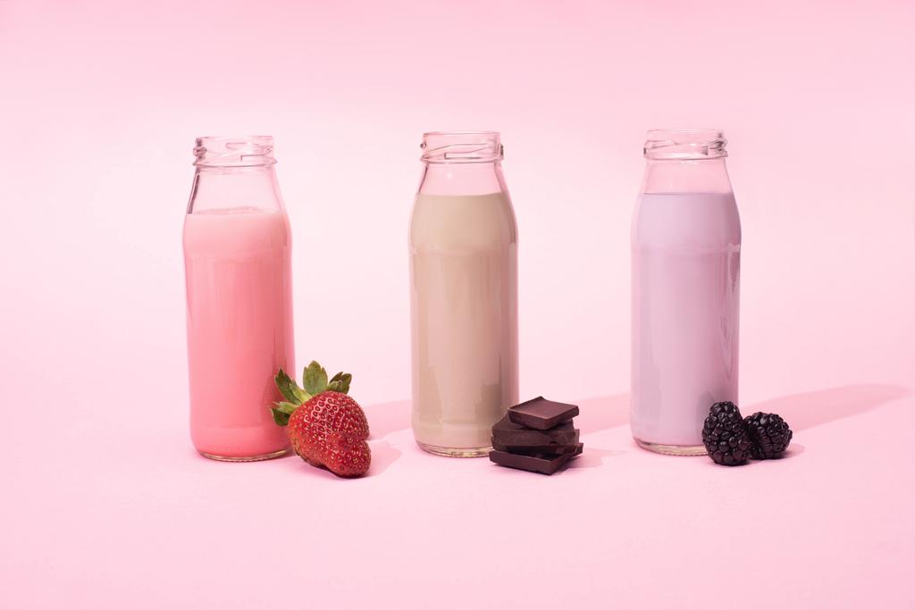 Bottles of milkshakes with strawberry, blackberries and pieces of chocolate on pink background - Photo, Image