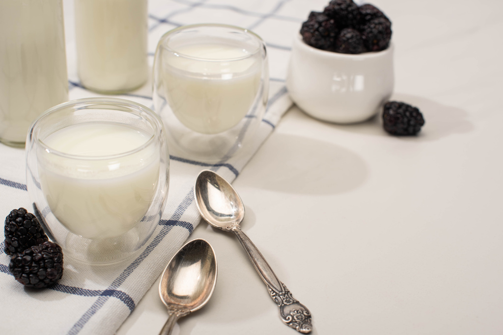 Focus selettivo di bottiglie e bicchieri di yogurt fatto in casa con more su panno vicino a cucchiaini su bianco
 - Foto, immagini
