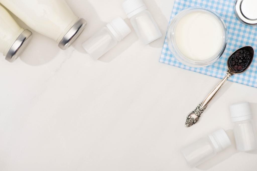 Vista superior de botellas y vaso de yogur, cucharadita con mora en tela cerca de contenedores con cultivos de iniciación en blanco
 - Foto, Imagen