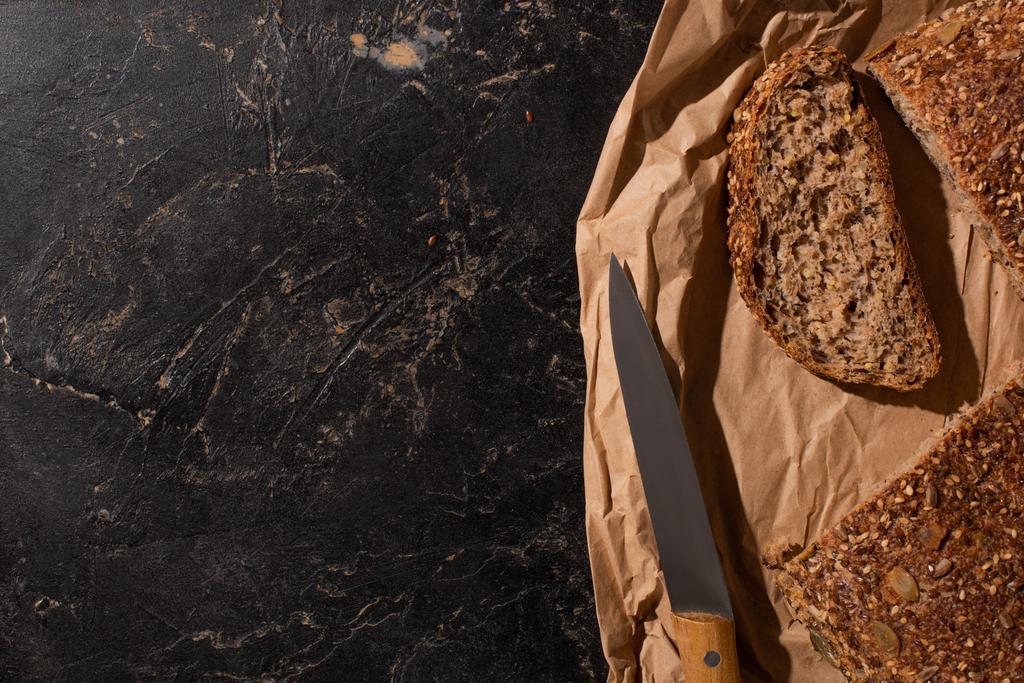 felső nézet vágott teljes kiőrlésű kenyér papíron közel kés kő fekete felületen - Fotó, kép