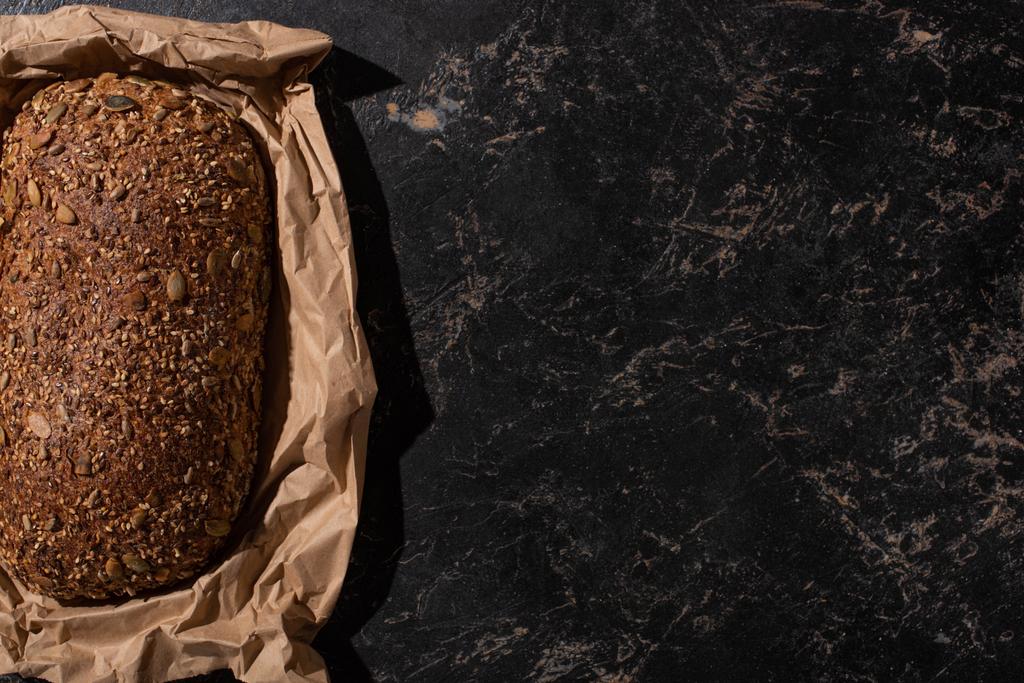top view of fresh baked loaf of whole grain bread in paper on stone black surface - Photo, Image
