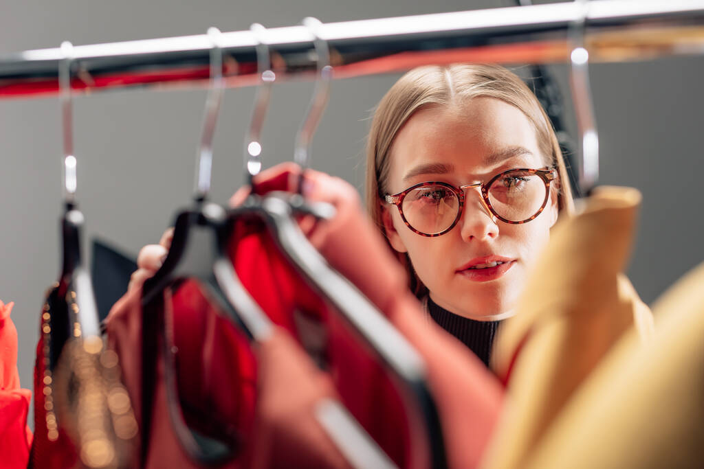 selektivní zaměření atraktivní stylistky v brýlích při pohledu na módní oblečení na ramínka  - Fotografie, Obrázek