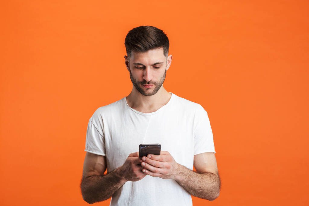 Beeld van een jonge man met een baard die basale kleding draagt en mobiele telefoon geïsoleerd houdt over oranje achtergrond - Foto, afbeelding