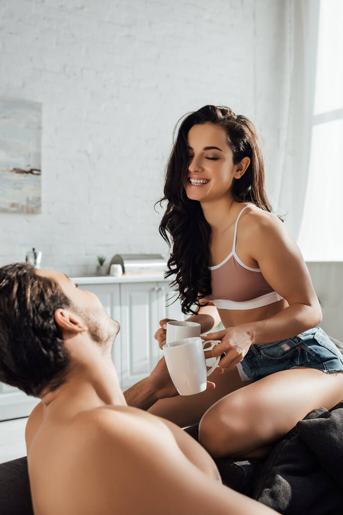 Mujer con los ojos cerrados sonriendo y dando taza de té a su novio en el sofá en la cocina
 - Foto, imagen