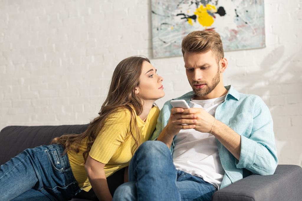 Attraktives Mädchen schaut abhängigen Freund mit Smartphone auf Couch an - Foto, Bild