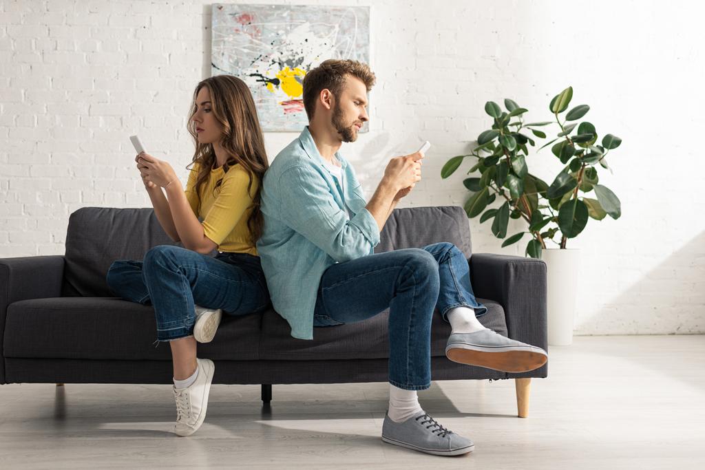 Seitenansicht eines Paares, das Smartphones benutzt, während es sich Rücken an Rücken auf dem Sofa zurücklehnt - Foto, Bild