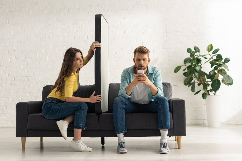 Νεαρή γυναίκα κρατώντας το μοντέλο του smartphone, ενώ ο φίλος κουβεντιάζοντας στον καναπέ  - Φωτογραφία, εικόνα