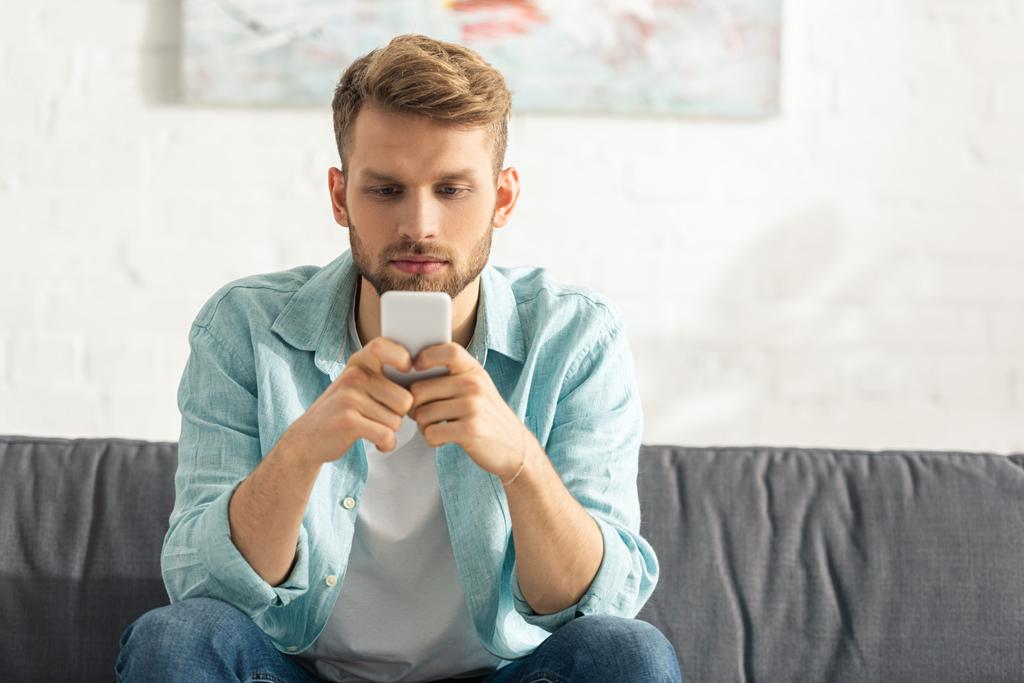 Schöner Mann mit Smartphone, während er zu Hause auf der Couch sitzt  - Foto, Bild
