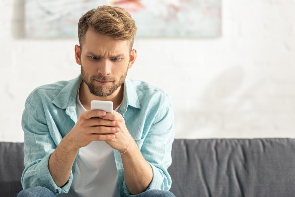 Homem confuso olhando para smartphone no sofá em casa
  - Foto, Imagem
