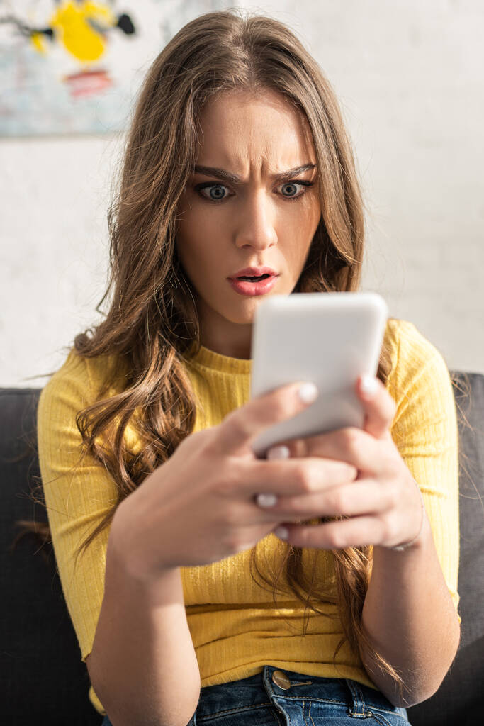 Selective focus of confused woman holding smartphone while sitting on couch  - Photo, Image