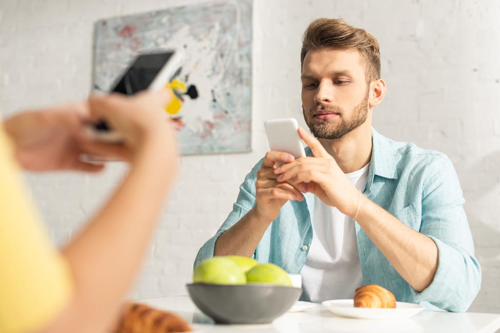 Επιλεκτική εστίαση του ανθρώπου χρησιμοποιώντας smartphone κοντά φίλη κατά τη διάρκεια του πρωινού στην κουζίνα  - Φωτογραφία, εικόνα