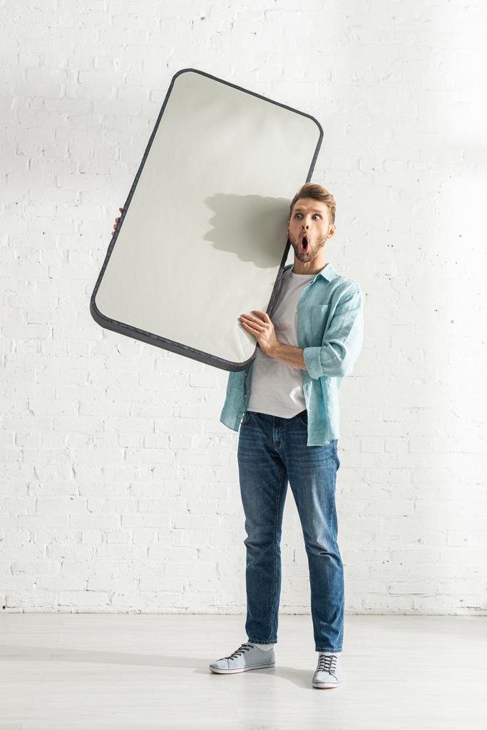 Zszokowany mężczyzna trzyma model smartfona w pobliżu białej cegły ściany  - Zdjęcie, obraz