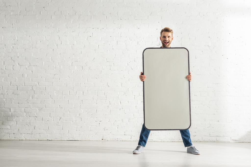 Vrolijke man met groot model van smartphone in de buurt van witte bakstenen muur  - Foto, afbeelding