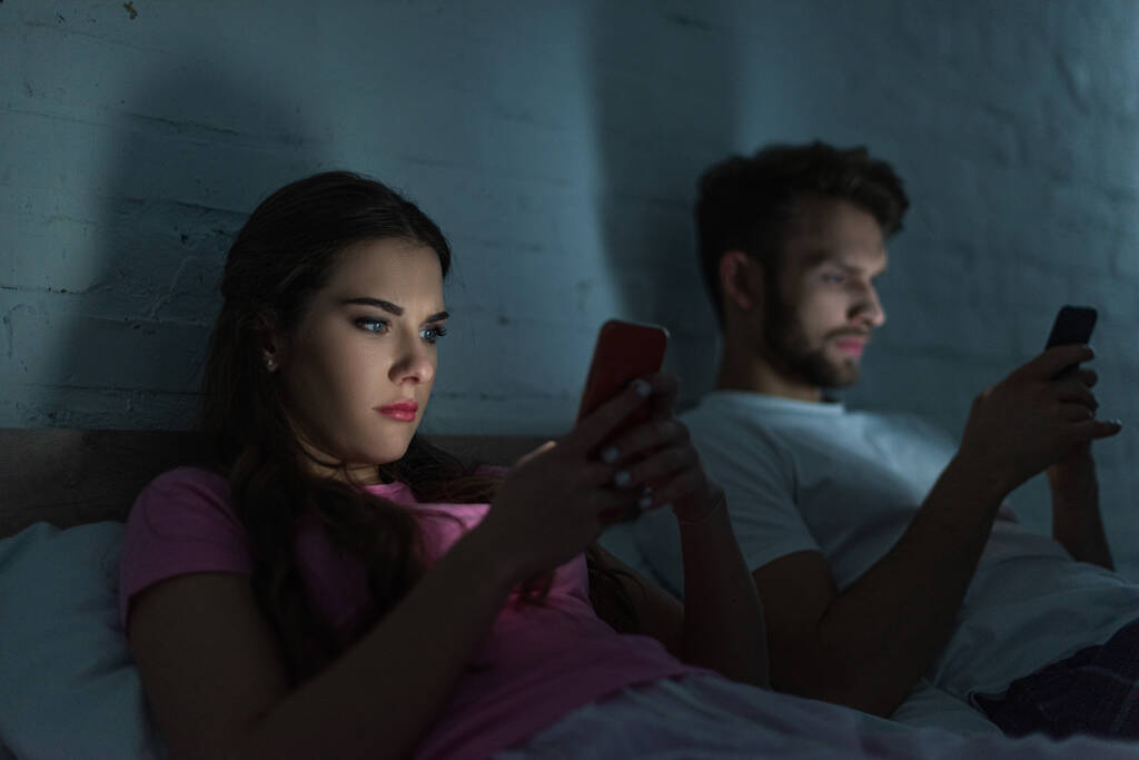 Selective focus of woman using smartphone near boyfriend chatting on bed at night  - Photo, Image