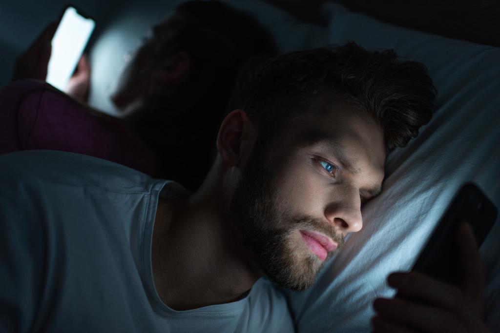 Enfoque selectivo del hombre guapo usando el teléfono inteligente cerca de la novia en la cama por la noche
  - Foto, imagen