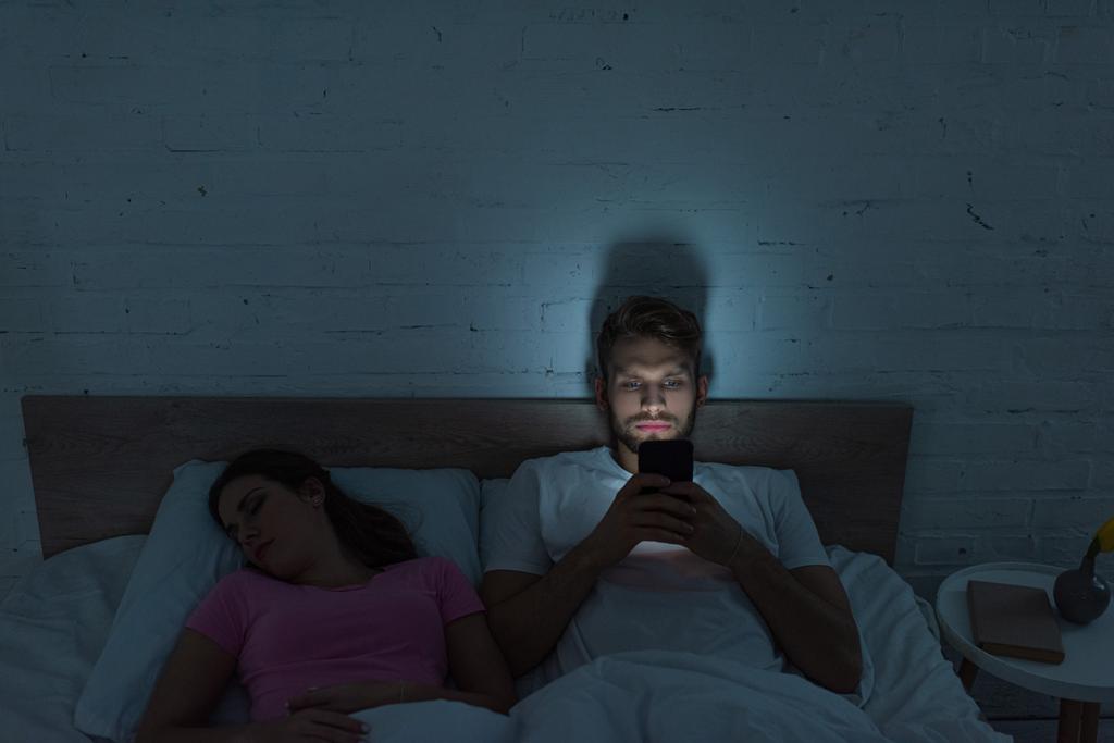 Смартфон зависит мужчина сидит на кровати рядом со спящей подругой
  - Фото, изображение