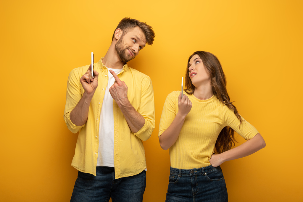Skeptical couple gesturing while showing smartphones at each other on yellow background - Photo, Image