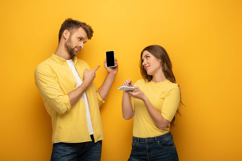 Pareja joven apuntando a teléfonos inteligentes mientras se miran el uno al otro en el fondo amarillo
 - Foto, Imagen