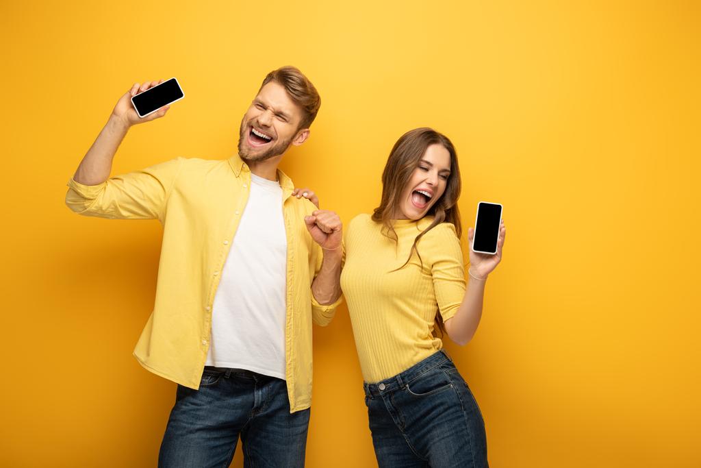 陽気若いですカップルは黄色の背景に空白の画面を持つスマートフォンを保持 - 写真・画像