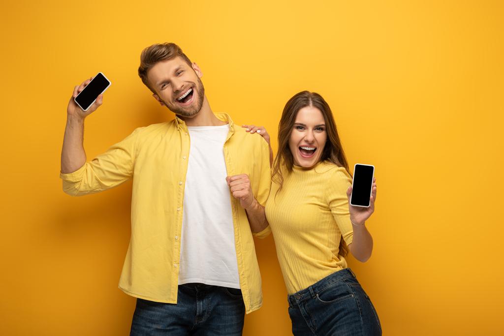 Jovem casal alegre mostrando smartphones e olhando para a câmera no fundo amarelo
 - Foto, Imagem
