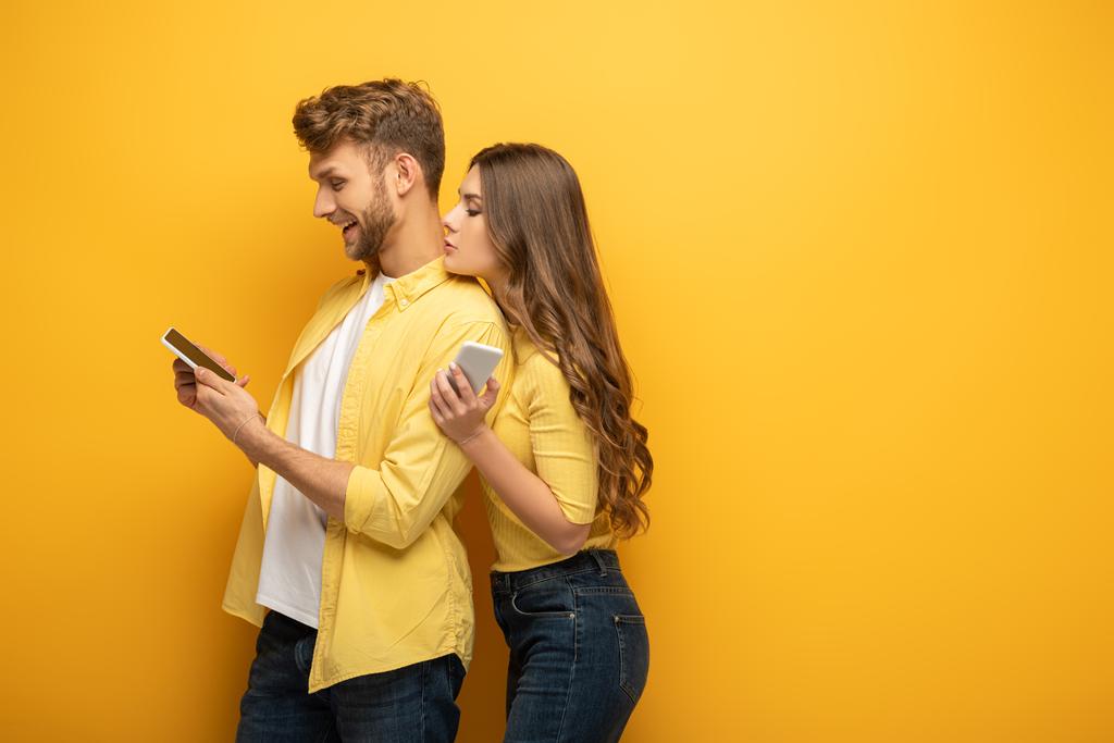 Vista lateral de pareja joven mirando el teléfono inteligente sobre fondo amarillo
 - Foto, Imagen