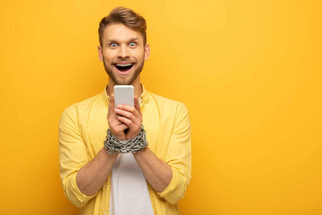 Cheerful man with metal chain around hands holding smartphone on yellow background - Photo, Image