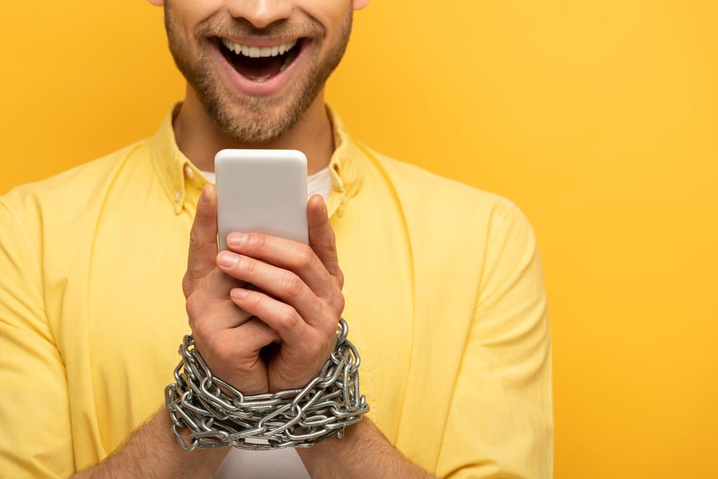 Vista recortada del hombre feliz con cadena alrededor de las manos sosteniendo el teléfono inteligente sobre fondo amarillo
 - Foto, Imagen