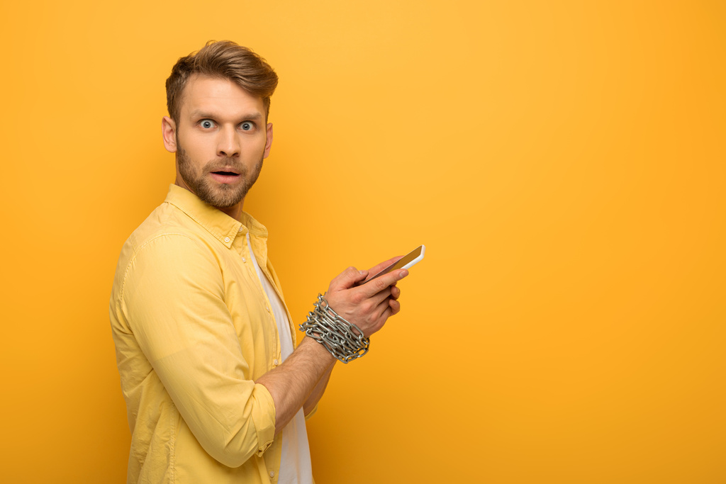 Side view of confused man with metal chain around hands holding smartphone and looking at camera on yellow background - Photo, Image
