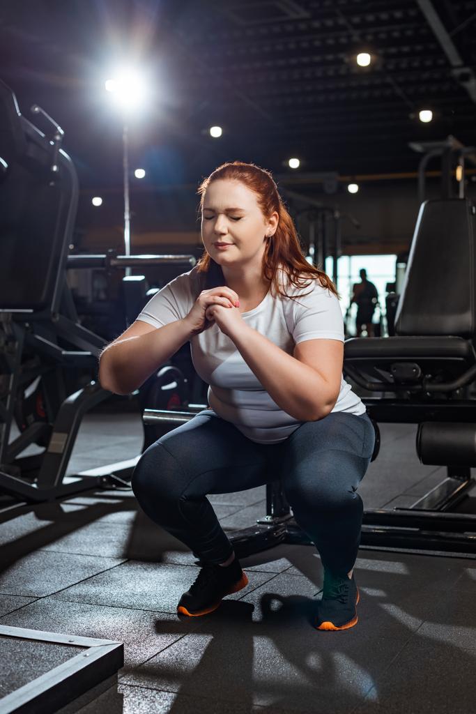 concentrated overweight girl with closed eyes squatting with clenched hands in gym - Photo, Image