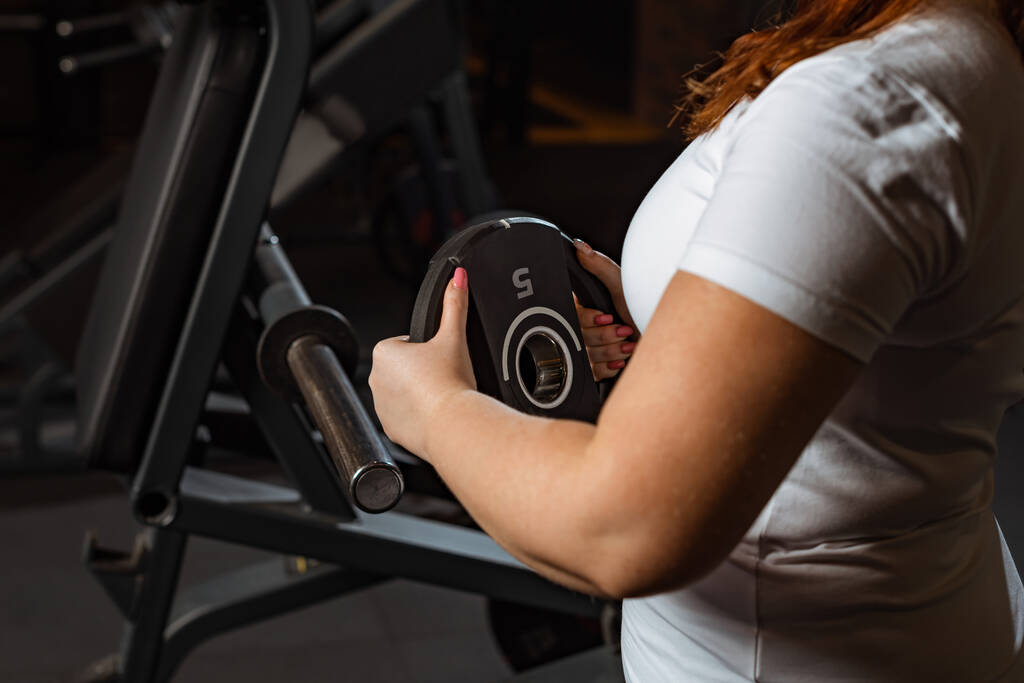 vista recortada de la chica con sobrepeso tomando el disco de peso de la máquina de fitness
 - Foto, imagen
