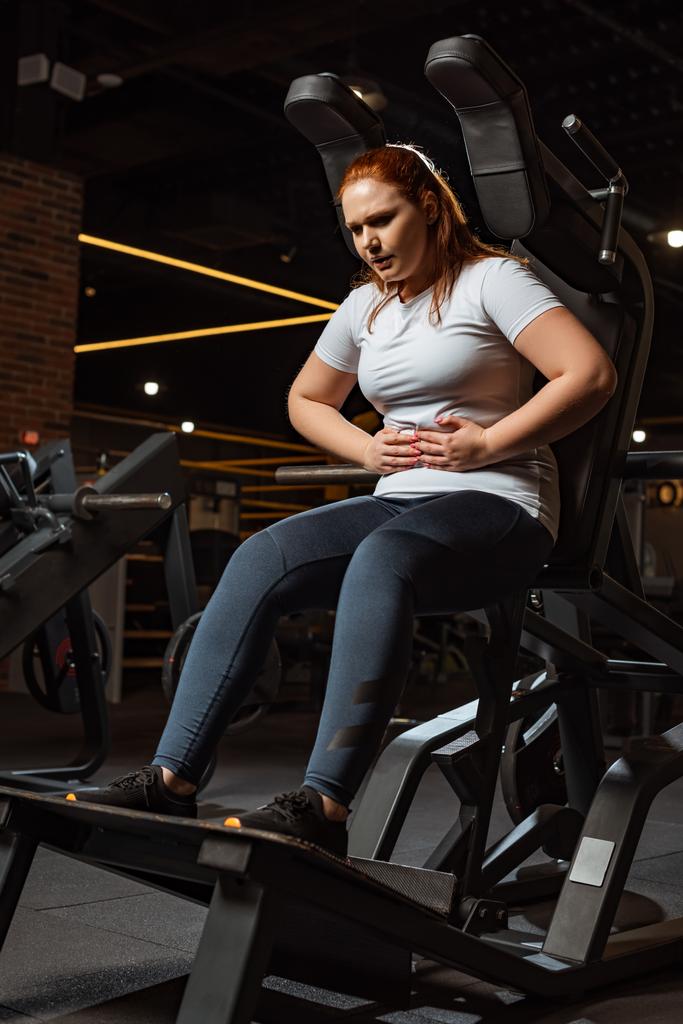 cansada chica con sobrepeso tocando el vientre mientras está sentado en la máquina de entrenamiento
 - Foto, Imagen