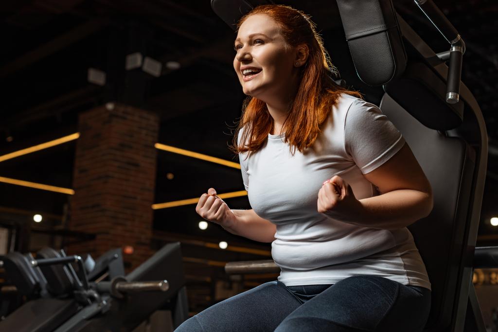 ενθουσιασμένοι υπέρβαρο κορίτσι δείχνει νικητής χειρονομία, ενώ κάθεται στο μηχάνημα γυμναστικής - Φωτογραφία, εικόνα