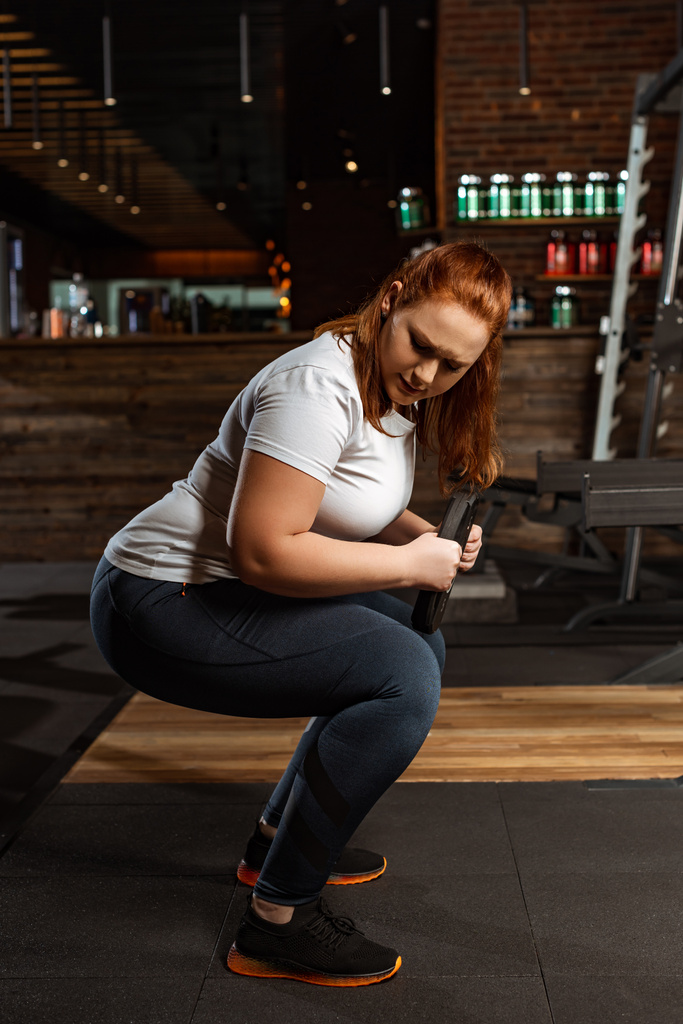 Konzentriertes übergewichtiges Mädchen hockt mit Gewichtsscheibe in Turnhalle - Foto, Bild