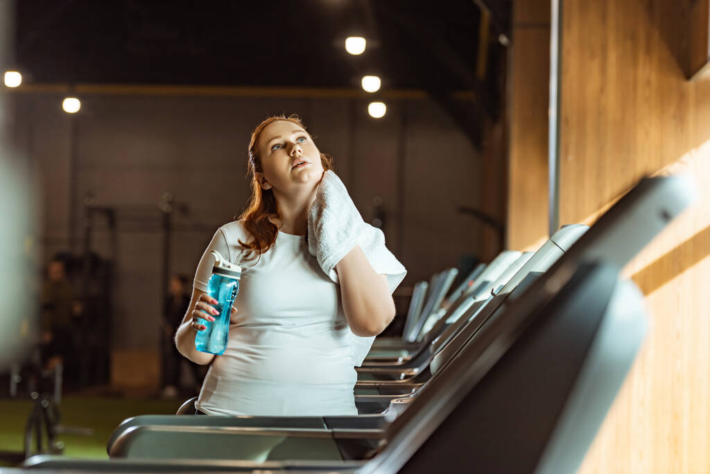 selective focus overweight girl wiping neck with towel while holding sports bottle  - Photo, Image